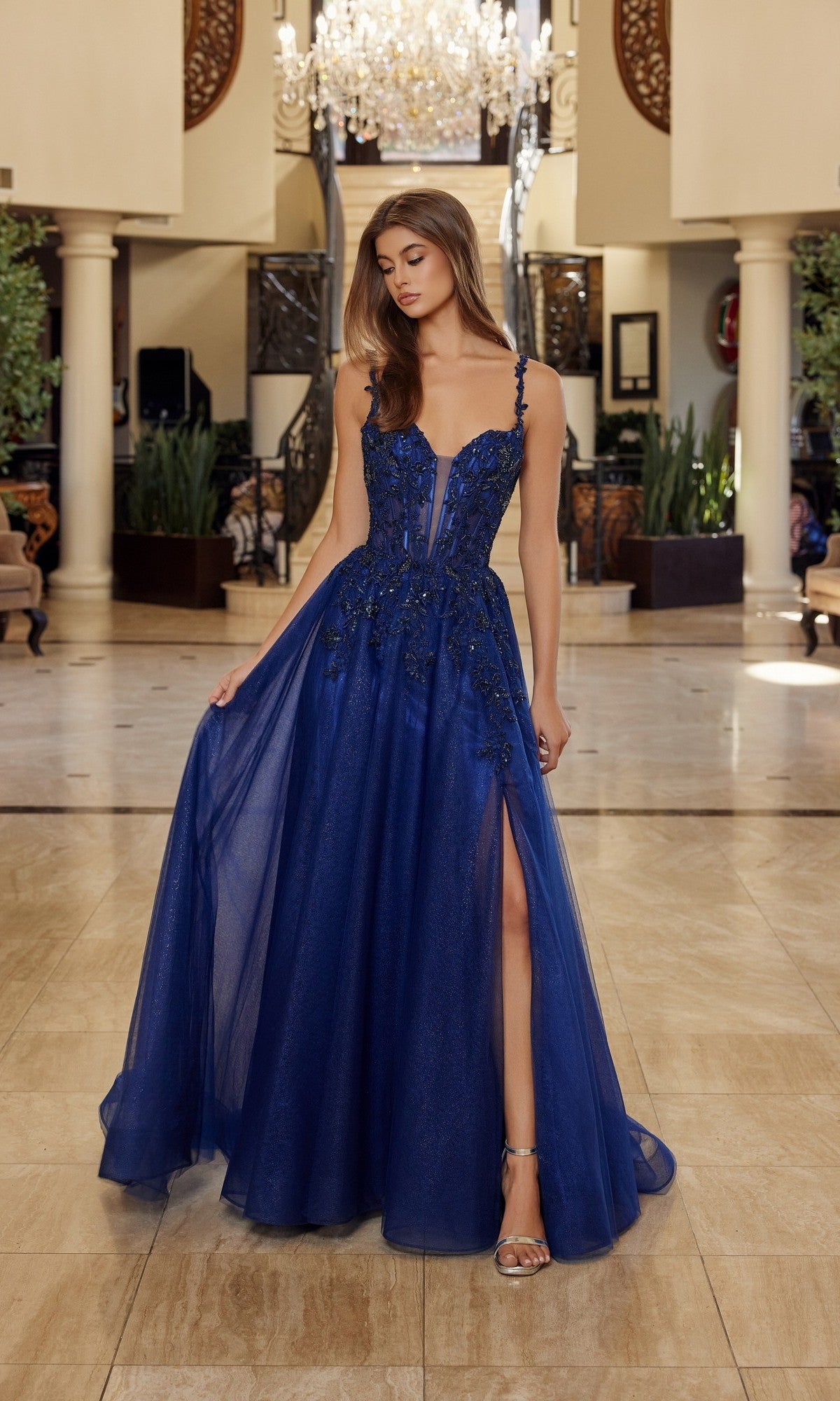 A woman in the Nox Anabel R1824 Long Prom Dress, navy blue with a floral embroidered corset, sheer overlay, and thigh-high slit, stands in an elegant lobby illuminated by chandeliers.